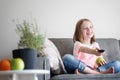 Adorable child girl 8 years old sits on a gray sofa with a green apple and a TV remote control in her hands rest and entertainment Royalty Free Stock Photo