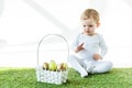 Child sitting on green grass near straw basket with traditional Easter eggs isolated on white Royalty Free Stock Photo