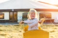 Adorable cute caucasian blond kid girl sitting in wooden cart having fun throwing straw or hay at farm or park during warm autumn