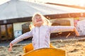 Adorable cute caucasian blond kid girl sitting in wooden cart having fun throwing straw or hay at farm or park during warm autumn