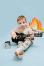 Adorable cute little boy sitting next to fake campfire, playing on little guitar