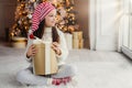 Adorable female child wears santa claus hat, warm clothes, hold wrapped gift box, sits against decorated New Year tree. Happy pret Royalty Free Stock Photo