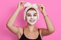 Adorable funny woman with clay mask on her face and hair band with bow on head touching her headband and looking at camera, has Royalty Free Stock Photo