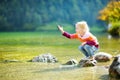 Adorable girl playing by Konigssee lake in Germany on warm summer day. Cute child having fun feeding ducks and throwing stones int