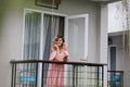 Adorable girl with wavy hair standing at balcony in morning. Glad blonde female model in pink dress