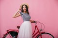 Adorable girl with wavy hair standing near red bicycle. Indoor photo of pleasant slim woman in hat