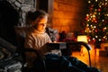 Adorable happy little blonde child girl opening magical Christmas box with gift sitting on rocking chair in xmas night Royalty Free Stock Photo