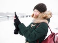 Adorable happy young redhead woman in green parka hat having fun at snowy winter exploring river pier doing photos on smartphone