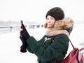 Adorable happy young redhead woman in green parka hat having fun at snowy winter exploring river pier doing photos on smartphone