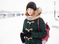 Adorable happy young redhead woman in green parka hat having fun at snowy winter exploring river pier doing photos on smartphone