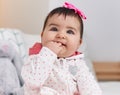 Adorable hispanic baby bitting fingers sitting on bed at bedroom