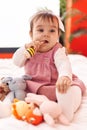 Adorable hispanic baby bitting maraca sitting on bed at bedroom