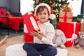 Adorable hispanic boy hearing gift sound sitting on floor by christmas tree at home