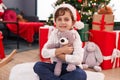 Adorable hispanic boy hugging teddy bear sitting on floor by christmas tree at home Royalty Free Stock Photo