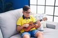 Adorable hispanic boy playing ukulele sitting on sofa at home Royalty Free Stock Photo