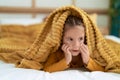 Adorable hispanic girl lying on bed covering with bedsheet at bedroom