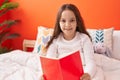 Adorable hispanic girl reading book sitting on bed at bedroom Royalty Free Stock Photo