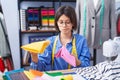 Adorable hispanic girl tailor holding cloths with doubt expression at clothing factory