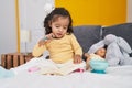 Adorable hispanic toddler sitting on bed reading book at bedroom Royalty Free Stock Photo