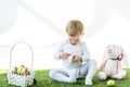 Kid holding white box while sitting on green grass near toy rabbit and straw basket with Easter eggs isolated on white Royalty Free Stock Photo