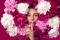 Adorable lady with fresh skin surrounded with peony flowers, keeping finger near lips, calls on to keep silence, woman with Royalty Free Stock Photo