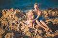 Adorable lady mother sitting on rocks beach in light blue dress together with her cute little blond daughter wearing pink airy dre Royalty Free Stock Photo