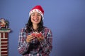 Adorable Latin girl smiling while holding a little Christmas present. wears a santa hat and lumberjack shirt. in the background