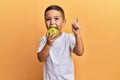 Adorable latin toddler smiling happy eating green apple looking to the camera over isolated yellow background