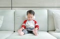 Adorable little Asian baby boy in headphones is using a smartphone lying on the sofa at home. Child listening to music on Royalty Free Stock Photo
