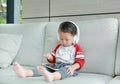 Adorable little Asian baby boy in headphones is using a smartphone lying on the sofa at home. Child listening to music on Royalty Free Stock Photo