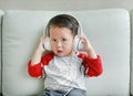 Adorable little Asian baby boy in headphones is using a smartphone lying on the sofa at home. Child listening to music on Royalty Free Stock Photo