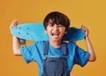 An adorable little asian boy looking happy while holding his skateboard against an orange background. Cute boy wearing