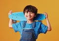 An adorable little asian boy looking happy while holding his skateboard against an orange background. Cute boy wearing