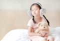 Adorable little Asian child girl wearing winter earmuffs and hugging teddy bear while sitting on the bed at home Royalty Free Stock Photo