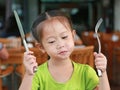 Adorable little Asian girl holding knife and fork sitting on wood chair at restaurant having breakfast Royalty Free Stock Photo