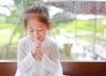 Adorable little Asian girl praying at glass windows on the raining day. Spirituality and religion Royalty Free Stock Photo