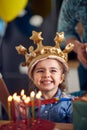 Adorable little birthday girl wearing inflatable golden crown, sitting in front of cake with lit candles, smiling feeling joyful