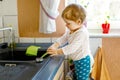 Adorable little blond baby girl washing dishes in domestic kitchen. Royalty Free Stock Photo