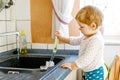 Adorable little blond baby girl washing dishes in domestic kitchen. Royalty Free Stock Photo