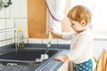 Adorable little blond baby girl washing dishes in domestic kitchen. Royalty Free Stock Photo