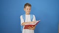 Adorable little blond boy student confidently smiling, joyfully reading a book while standing against an isolated blue background Royalty Free Stock Photo