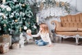 Adorable little blonde girl in a blue jeans sitting on a floor near christmas tree and looking on toy. Royalty Free Stock Photo