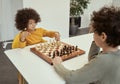 Adorable little boy with afro hair making a move while playing chess with his friend, sitting together at the table Royalty Free Stock Photo