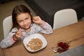 Adorable little child girl cutely smiling looking at camera, sitting at table and taking her delicious healthy breakfast Royalty Free Stock Photo