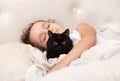 Adorable little cute girl child is sleeping in the bed with her pet black cat. The girl is hugging the teddy bear Royalty Free Stock Photo