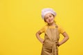 Adorable little girl in chef's apron and hat, with hands on waist, smiles looking at camera, isolated yellow Royalty Free Stock Photo