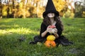 Adorable little girl with hair in black witch hat making grimace, holding pumpkins, on a sunny autumn day background.