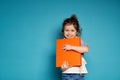 Adorable little girl gently hugs an orange book and cute smiles with toothy smile looking at camera while standing on blue