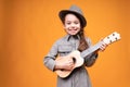 Adorable little girl in a gray hat smiles and plays the guitar while standing on an orange background