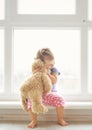 Adorable little girl hugging a teddy bear. Cute baby at home in white room is sitting near window. Royalty Free Stock Photo